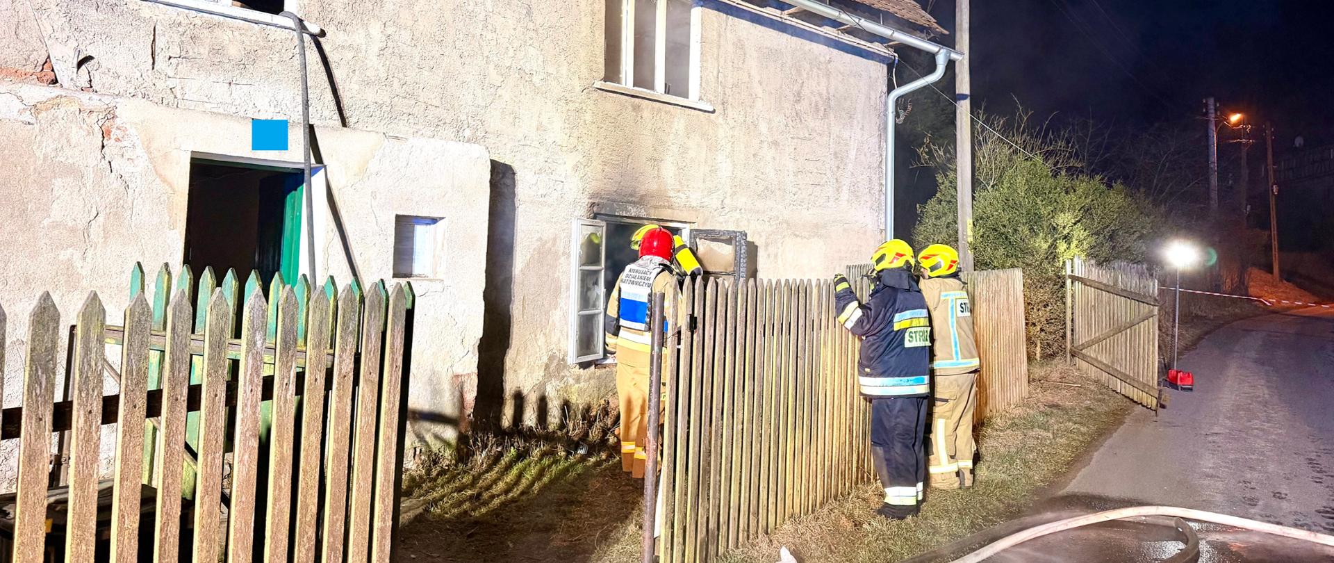 Budynek jednorodzinny, przy otwartym oknie na parterze stoją strażacy, okolice okna okopcone