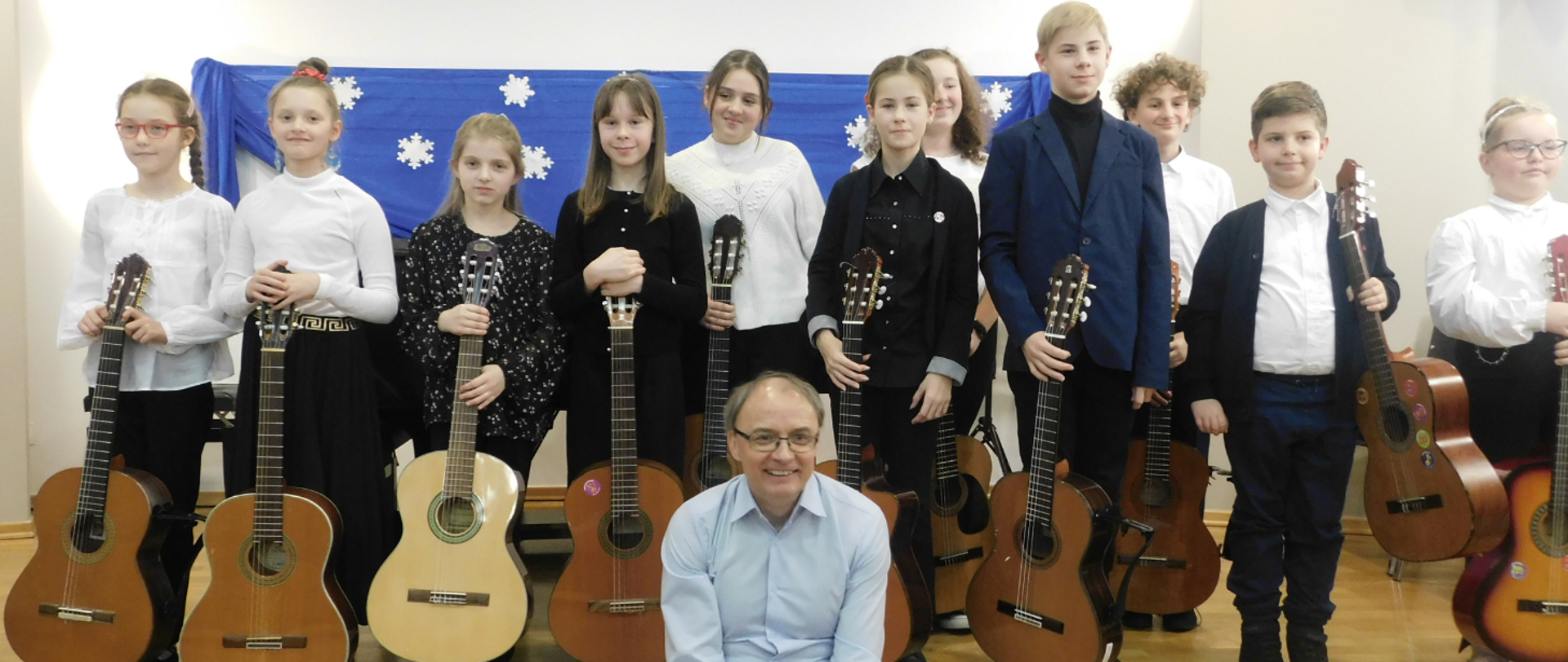 Na zdjęciu grupa odświętnie ubranych uczniów, którzy trzymają gitary. Przed nimi siedzi uśmiechnięty mężczyzna w okularach, który ubrany jest w błękitną koszulę.