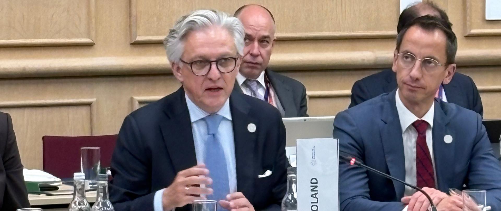 Uczestnicy oficjalnego spotkania siedzą przy stole konferencyjnym. Osoba przy tabliczce z napisem „Denmark” zabiera głos, a osoba siedząca obok tabliczki „Poland” słucha wystąpienia. Na stole znajdują się mikrofony, dokumenty, notatniki, szklanki oraz karafki z wodą.