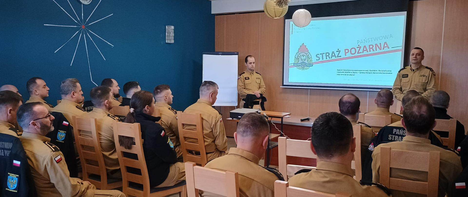 Dwóch funkcjonariuszy PSP prowadzi szkolenie podczas odprawy w Turawie. Na ekranie widnieje logo Państwowej Straży Pożarnej oraz tekst o znaczeniu szkolenia dla rozwoju kadry dowódczej. Uczestnicy siedzą w sali z niebieską ścianą i zegarem.