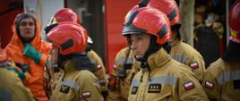 strażacy ubrani w kombinezony specjalne, na głowach mają czerwony hełmy, po lewej w oddali jeden strażak ubrany jest w pomarańczowe ubranie ochrony chemicznej