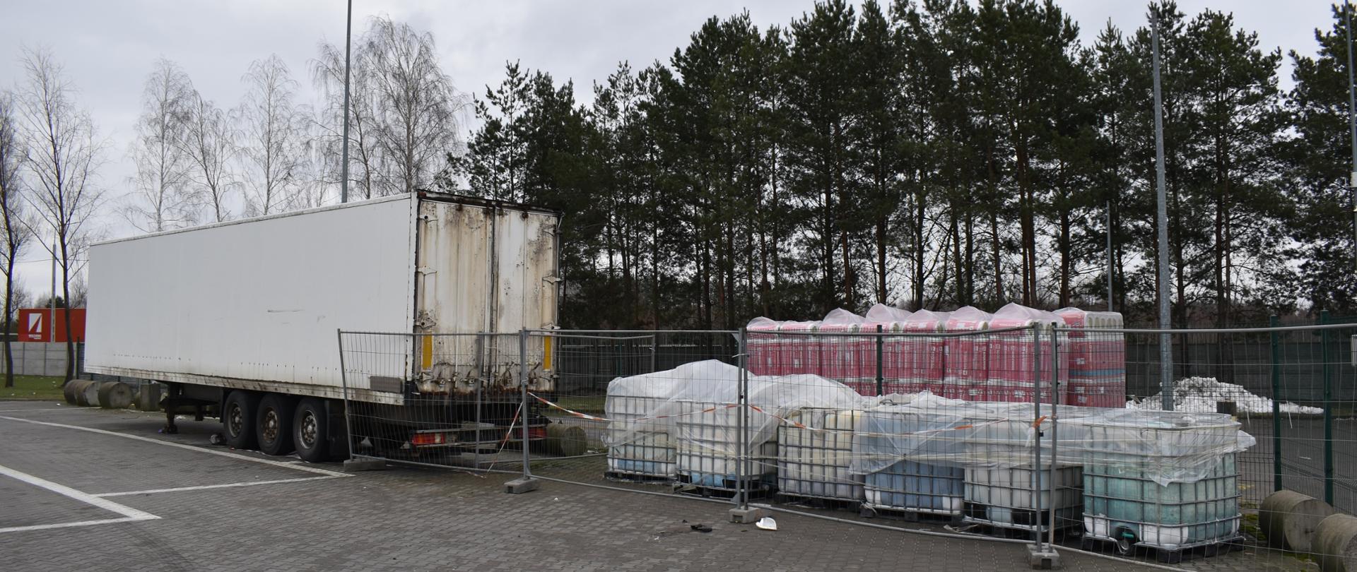 Naczepa samochodu ciężarowego oraz kilkanaście pojemników o pojemności 1000 l stoją na otwartym terenie.