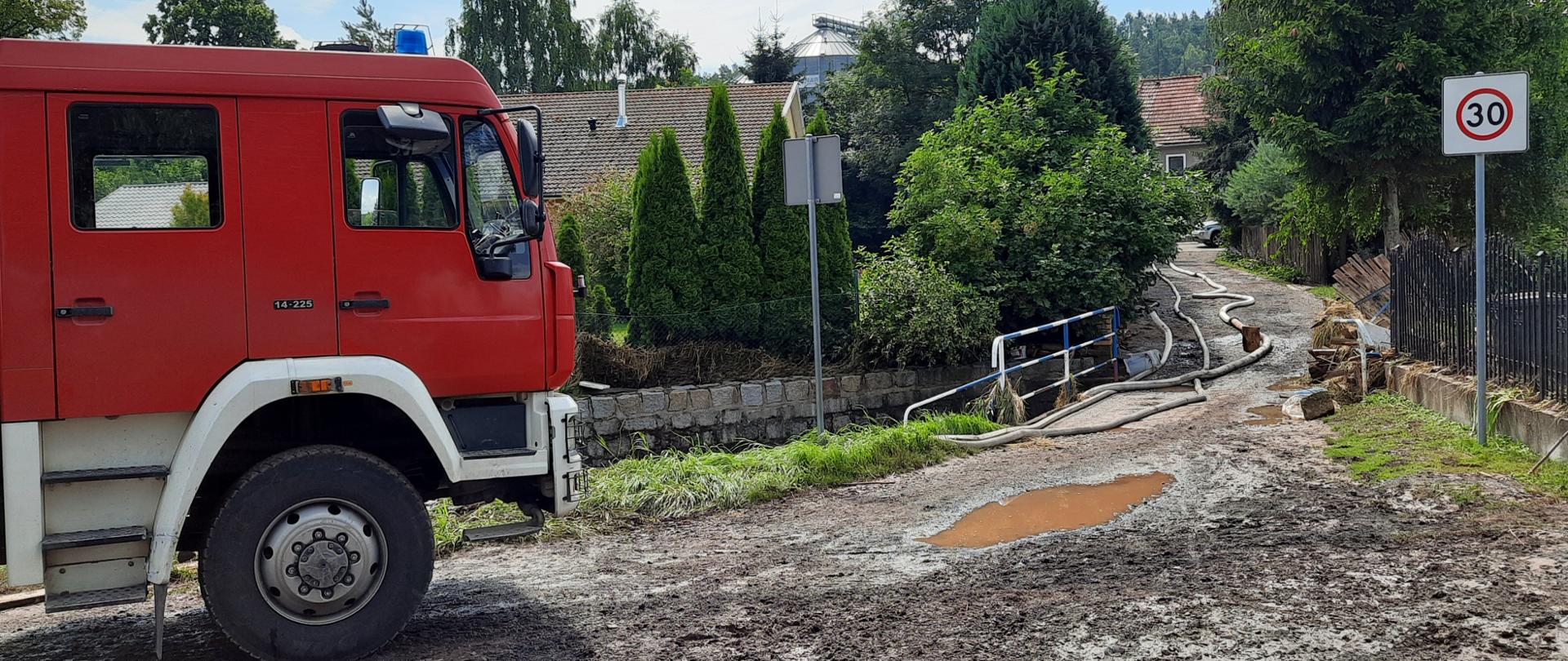 Zdjęcie przedstawia samochód pożarniczy z Jednostki Ratowniczo-Gaśniczej Państwowej Straży Pożarnej w Lwówku Śląskim na drodze dojazdowej do posesji. Widoczna zabłocona droga w wyniku wystąpienia wód ze strumienia. 