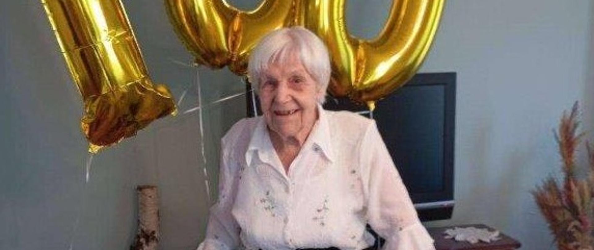 Uśmiechnięta starsza kobieta z krótkimi siwymi włosami w białej bluzce i czarno-białej spódnicy stoi przy chodziku ortopedycznym w pokoju, a za nią unoszą się złote balony w kształcie liczby 100. Z tyłu szafka, a na niej telewizor.