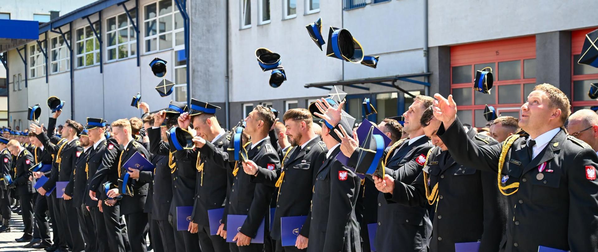 Zdjęcie przedstawia funkcjonariuszy Państwowej Straży Pożarnej, którzy w trakcie uroczystości promocji na wyższy stopień służbowych w geście celebracji podrzucają swoje czapki rogatywki.