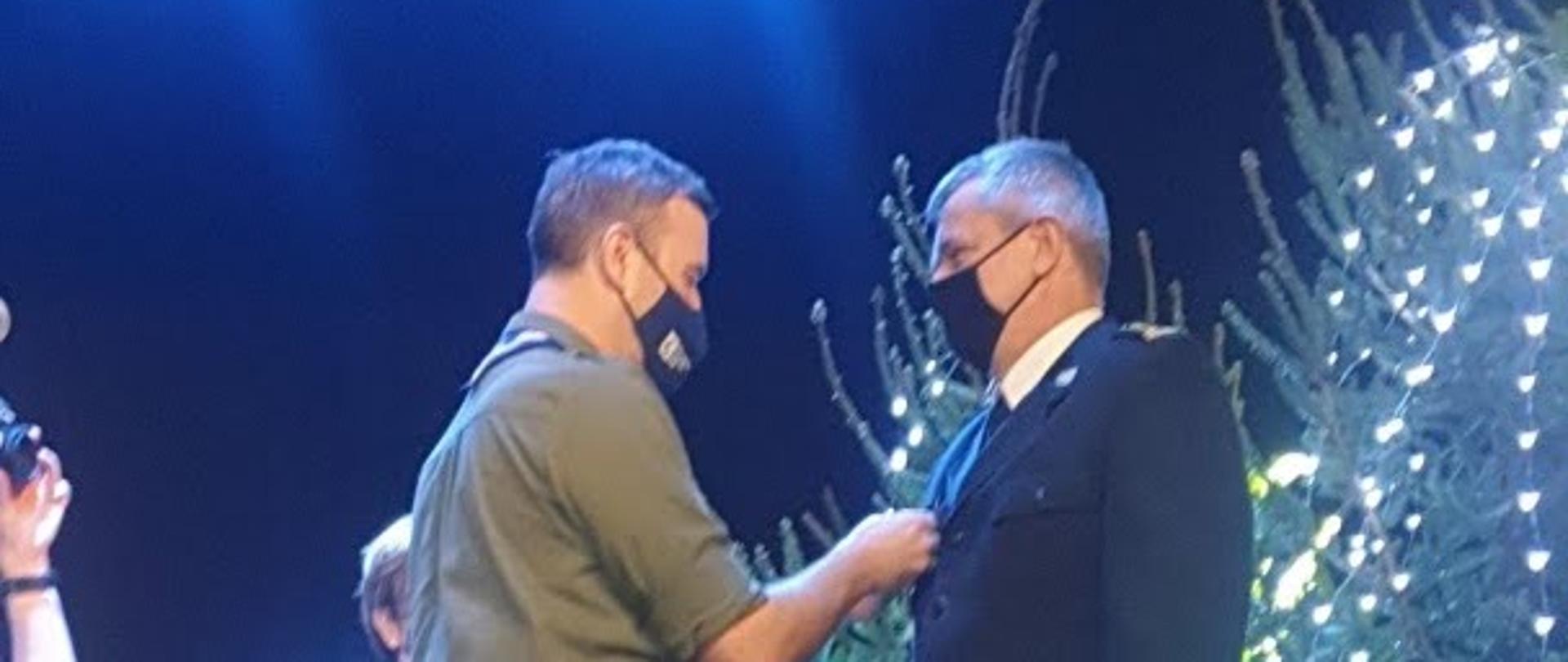 Na tle przystrojonych choinek komendant Chorągwi Gdańskiej Związku Harcerstwa Polskiego przypina pomorskiemu komendantowi wojewódzkiemu Państwowej Straży Pożarnej medal. Obok stoi harcerka z tacą.