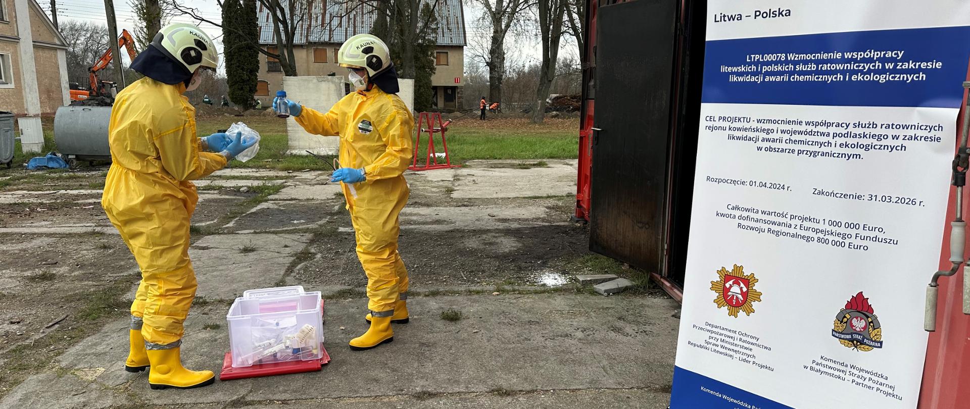 Szkolenia w ramach projektu LTPL00078 "Wzmocnienie współpracy litewskich i polskich służb ratowniczych w zakresie likwidacji awarii chemicznych i ekologicznych" - strażacy w żółtym ubraniu specjalnym w trakcie szkolenia, z boku po prawej stronie plakat projektowy 