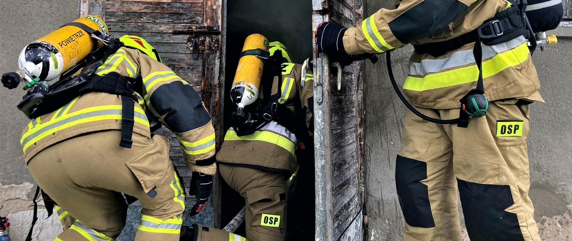 Trzech strażaków podczas ćwiczeń w trakcie wejścia do zadymionego budynku. Strażacy ubrani w ubrania specjalne, aparaty ochrony układu oddechowego oraz hełmy strażackie.