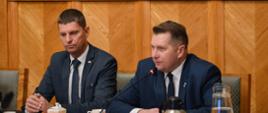 Przy stole siedzi minister Czarnek i wiceminister Piontkowski, minister mówi do mikrofonu. Za nimi ściana w zielony wzorek.