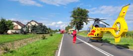 Słoneczny dzień na drodze na jednym pasie stoi żółty helikopter Lotniczego Pogotowia Ratunkowego, widać go z lewego boku oraz tylną część. Obok stoi osoba odwrócona placami ubrana w czerwony kombinezon z napisem na plecach Lotnicze Pogotowie ratunkowe. W oddali widać na poboczy srebrny samochód osobowy oraz dwa czerwone samochody Straży Pożarnej. Na środku jezdni stoi żółta karetka pogotowia.