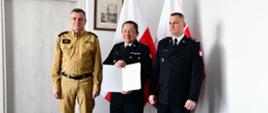 Pomorski komendant wojewódzki Państwowej Straży Pożarnej oraz strażacy stoją na tle flagi Rzeczypospolitej Polskiej. Jeden z funkcjonariuszy, będący w środku, trzyma list gratulacyjny.