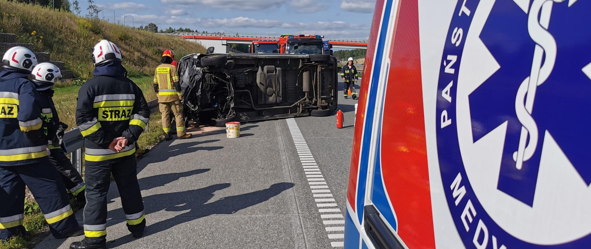 Woźniki - wypadek samochodu dostawczego - po lewej stronie 3 strażaków na drodze, po prawej stronie samochód ratownictwa medycznego, w tle kilku strażaków przy przewróconym na bok samochodzie dostawczym