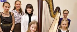 Na zdjęciu widać nauczycielkę i pięć dziewczynek. Stoją na tle jasnej ściany, pomiędzy nimi znajduje się harfa, jedna z dziewczynek przytula głowę do instrumentu.