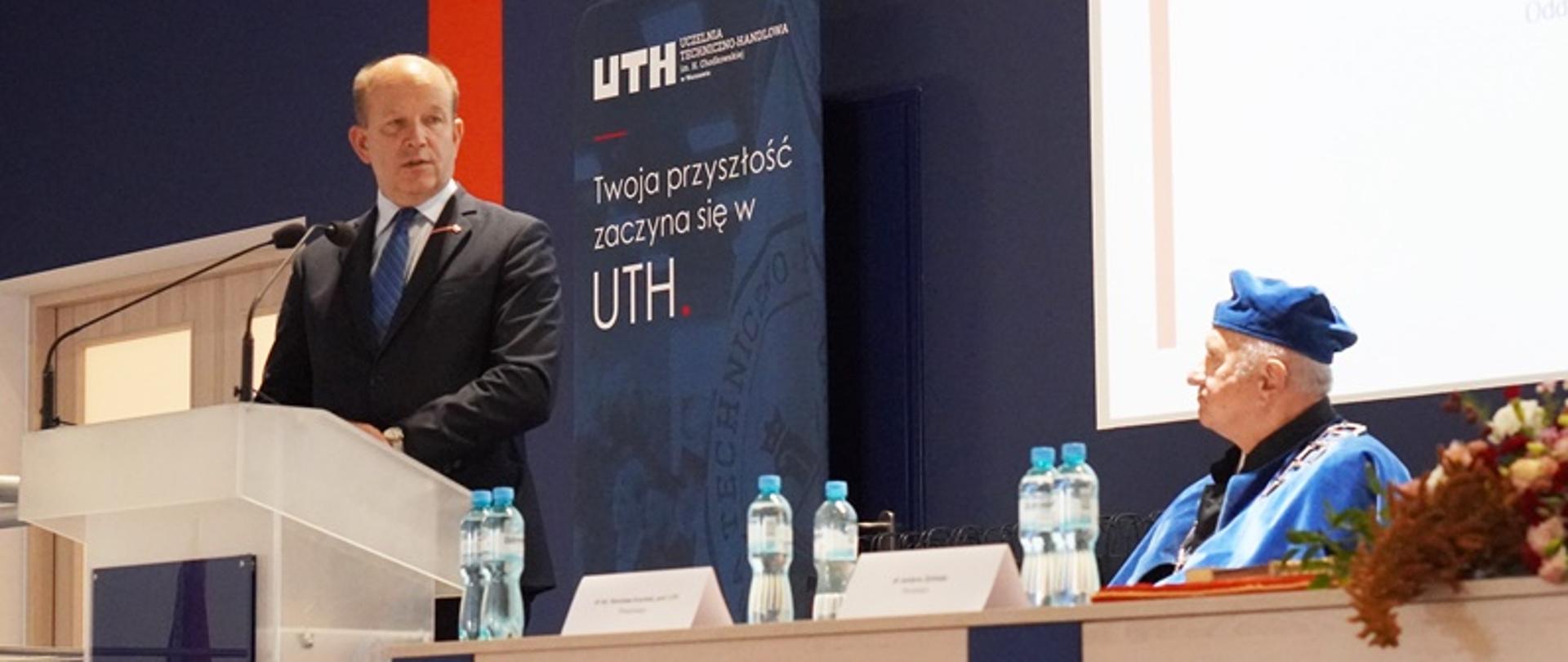 Na fotografii znajduje się dwóch mężczyzn, pierwszy z nich stoi przed mównicą z logo Uczelni Techniczno-Handlowej i jest ubrany w ciemnogranatową marynarkę, białą koszulę i niebieski krawat. Tuż obok mównicy znajduje się stół prezydialny, za którym widać ubranego w togę i biret prodziekan ds. studenckich i organizacyjnych uczelni. W tle ścianka wizerunkowa z napisem "Twoja przyszłość zaczyna się w UTH" oraz z logo uczelni