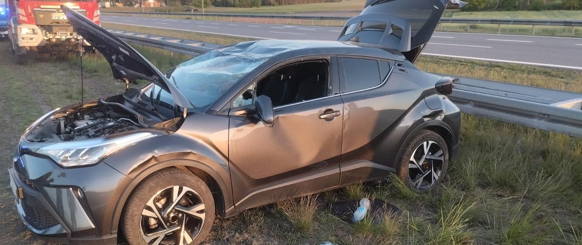 Na zdjęciu widać szarego crossovera marki Toyota, który uległ wypadkowi drogowemu. Samochód znajduje się na poboczu autostrady częściowo w trawie. Pojazd ma otwartą maskę i bagażnik, a jego karoseria jest widocznie uszkodzona — szczególnie po lewej stronie. Przednia szyba i boczne szyby są rozbite, a wokół samochodu na ziemi leży rozbita szyba oraz różne przedmioty. W tle widoczny jest wóz strażacki i część zabezpieczonej sceny wypadku.