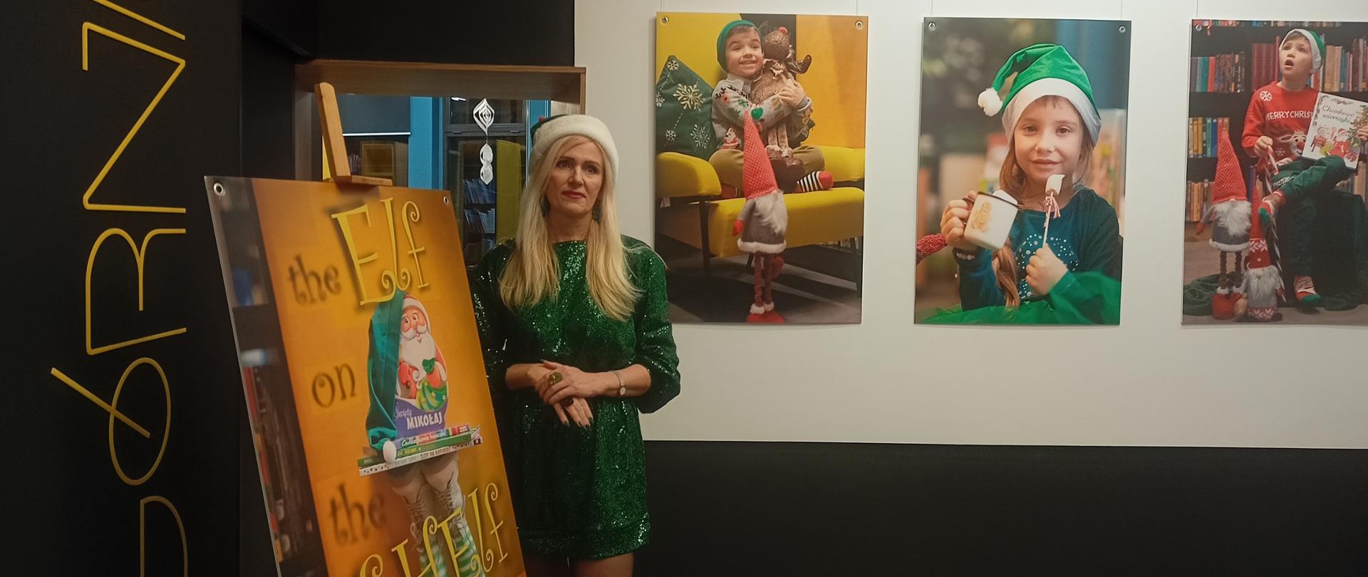 Kobieta ubrana w zielony strój elfa stoi przed plakatem z hasłem "Elf in the Shelf"