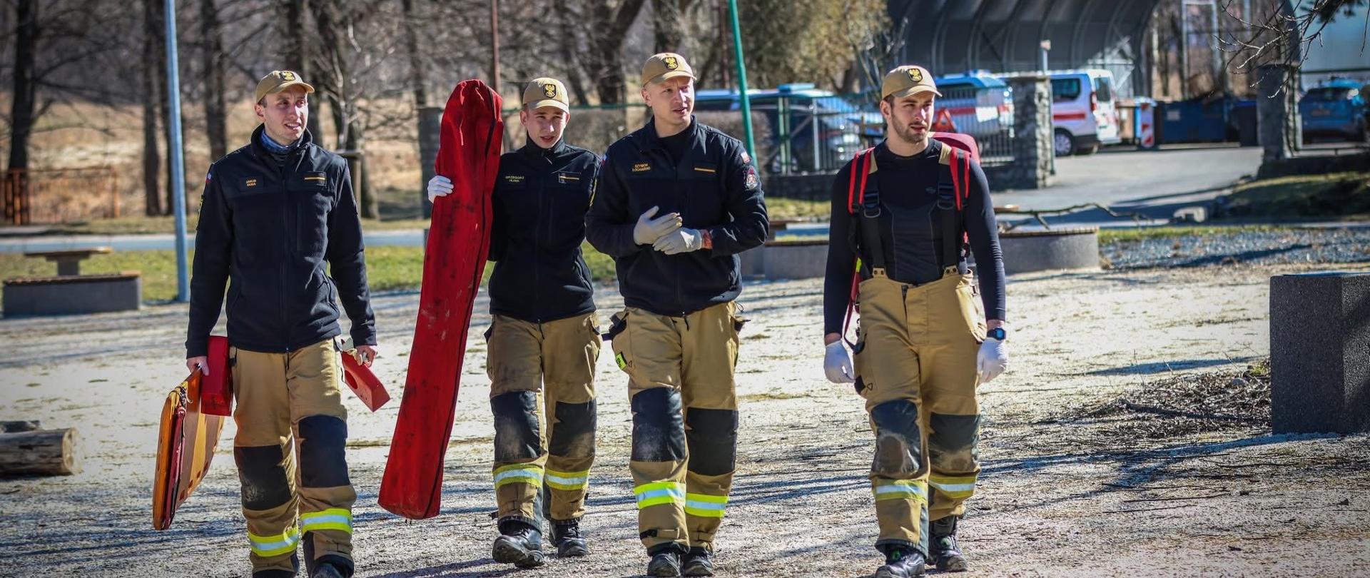 Zdjęcie przedstawia czterech strażaków idących obok siebie po placu na świeżym powietrzu. Wszyscy mają na sobie piaskowe, specjalistyczne spodnie bojowe z odblaskowymi pasami oraz ciemne bluzy. Trzech z nich niesie w dłoniach elementy wyposażenia ratowniczego, w tym długą, podłużną czerwoną torbę i deskę ortopedyczną. W tle, za idącymi mężczyznami, widoczne są drzewa bez liści, fragmenty zaparkowanych samochodów oraz zadaszona, półkolista konstrukcja (wiata).