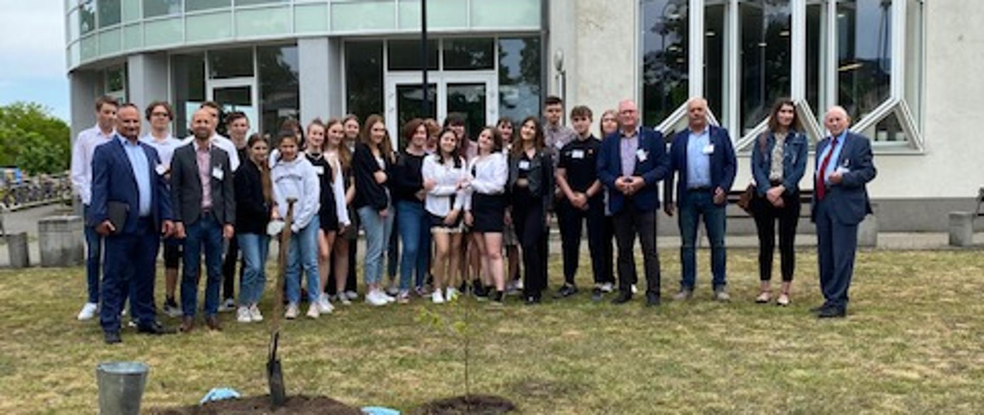 Grupa kilkunastu osób pozuje przed budynkiem. Przed ludzi w ziemi stoi niska sadzonka drzewa.