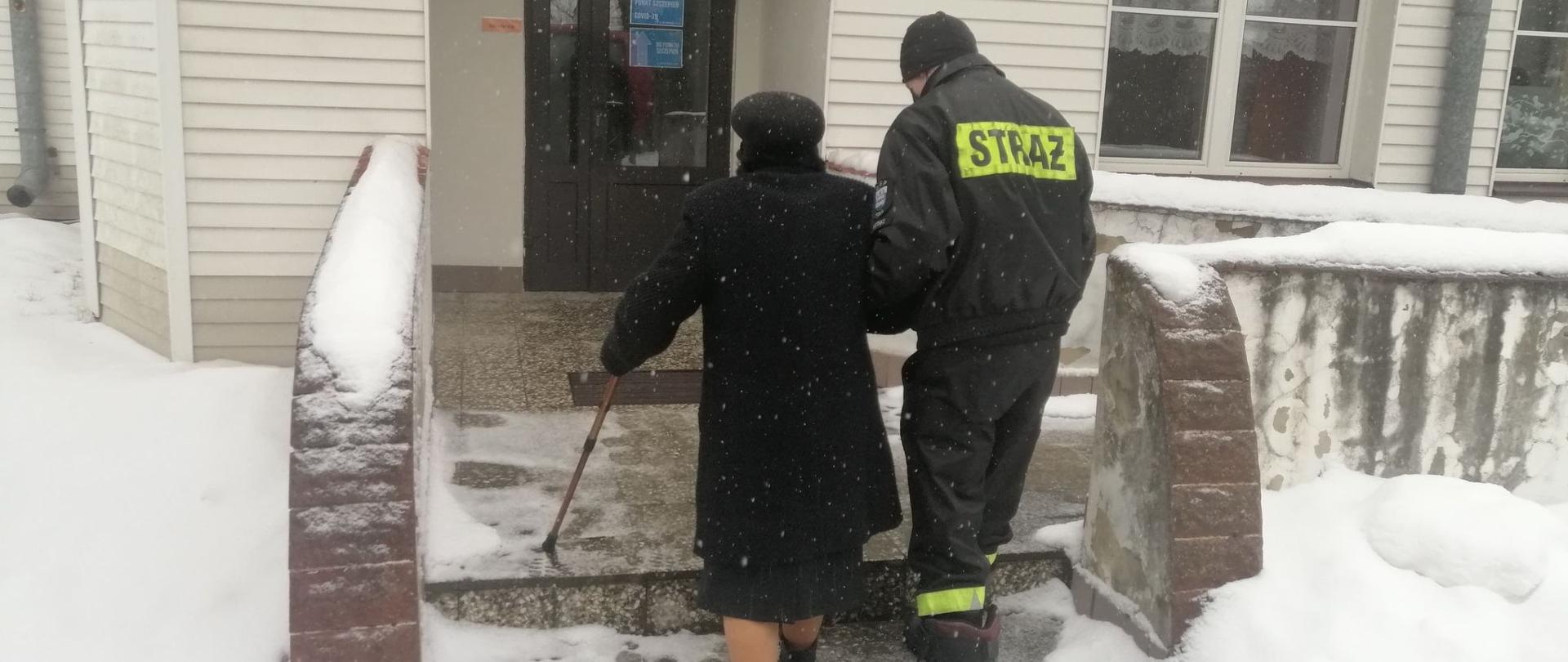 Strażak prowadzi starszą kobietę z laską po schodach do budynku przychodni