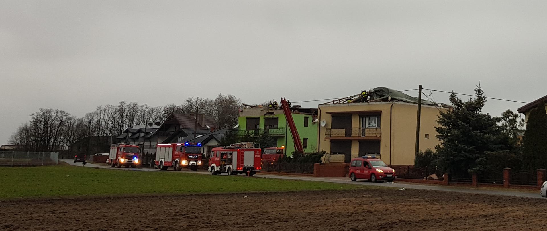 Na zdjęciu dwa budynki z zerwanymi całkowicie dachami. Samochody pożarnicze i strażacy w trakcie akcji.