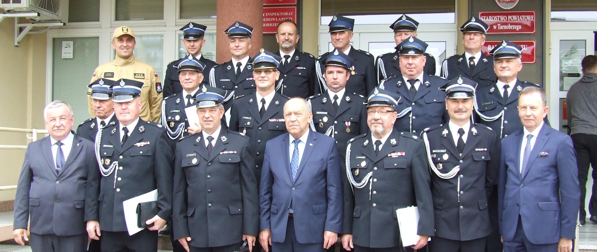 Na schodach przed wejściem do budynku, w trzech rządach stoją strażacy w mundurach. Wśród nich, w pierwszym rzędzie stoi również trzech mężczyzn w garniturach. 