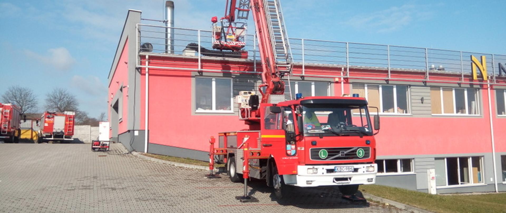 Zdjęcie przedstawia drabinę hydrauliczną, która jest rozłożona obok zakładu stolarskiego. W górnej części zdjęcia widzimy strażaków pracujących na dachu zakładu. Strażacy prowadzą wentylację zakładu przy pomocy elektrycznego wentylatora oddymiającego. Jest pora dzienna.