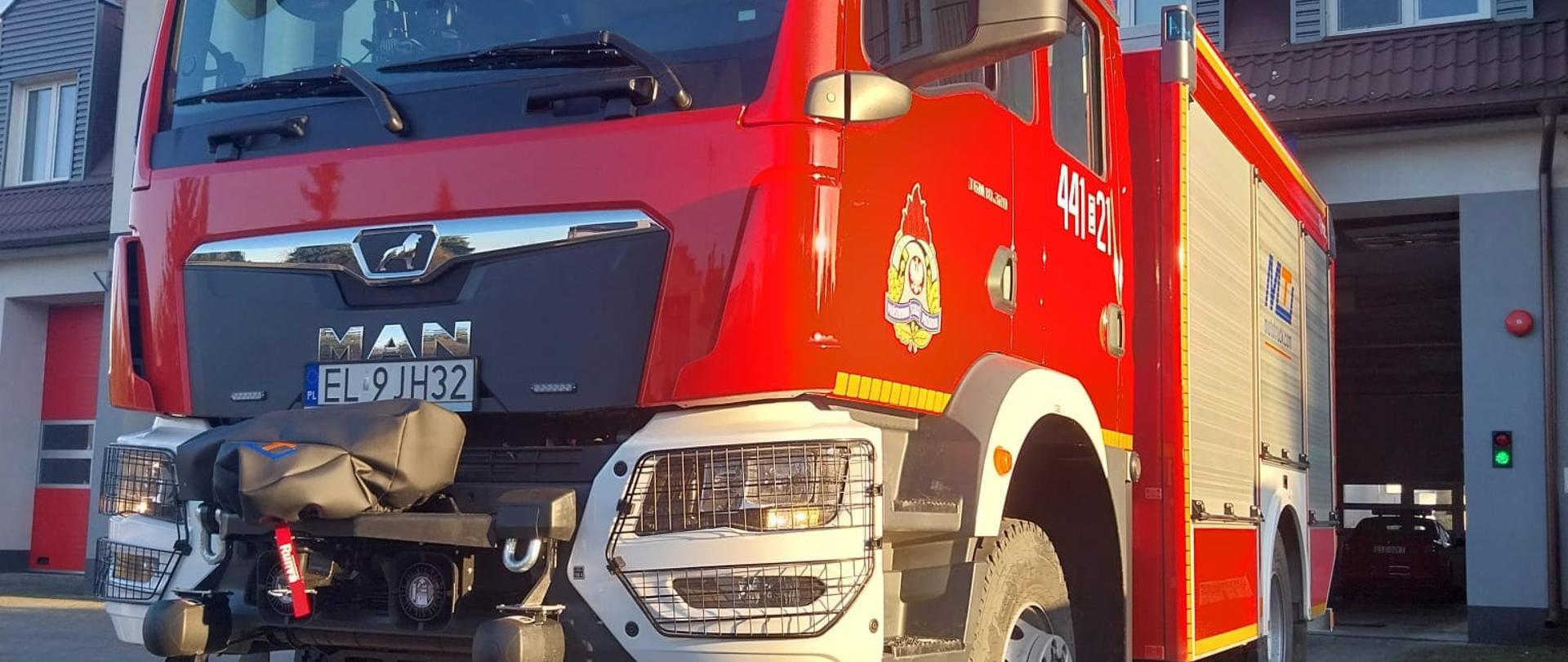Na zdjęciu widać duży czerwony samochód strażacki, który stoi przed budynkiem straży pożarnej. Na drzwiach ma logo Państwowej Straży Pożarnej i biały napis 441E21. Z przodu ma napis MAN i numer rejestracyjny EL9JH32. Pojazd ma zapalone światła. Jest widno.