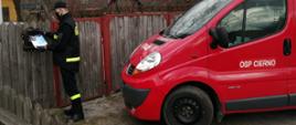 - Na pierwszym planie widnieje przód samochodu operacyjnego, strażackiego w kolorze czerwonym. Na drzwiach kierowcy widnieje napis „OSP Cierno-Żabieniec”. Po lewej stronie znajduje się strażak w umundurowaniu koszarowym z założoną maseczką ochronną oraz rękawiczkami. Strażak wrzuca ulotkę informacyjna do skrzynki na listy, która znajduje się płocie jednego z domostw.
