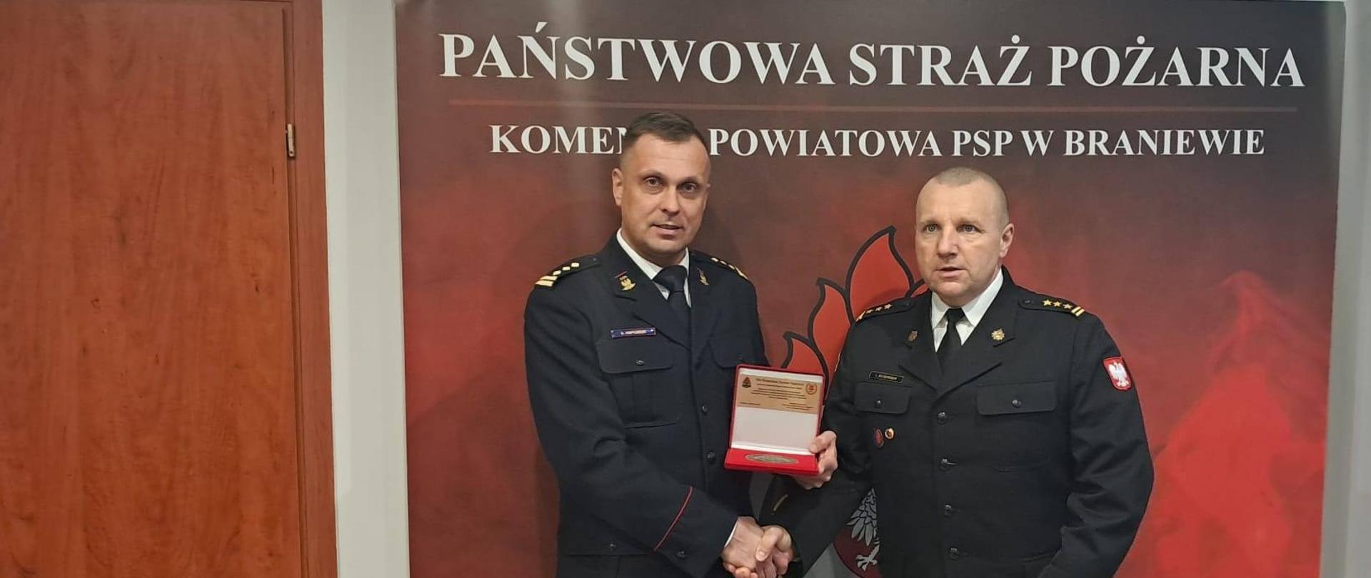 Na zdjęciu Komendant powiatowy Państwowej straży pożarnej z prawej i komendant służby ochrony kolei w mundurach wyjściowych. W tle ścinaka z napisem Państwowa Straż Pożarna Komenda Powiatowa PSP w Braniewie.