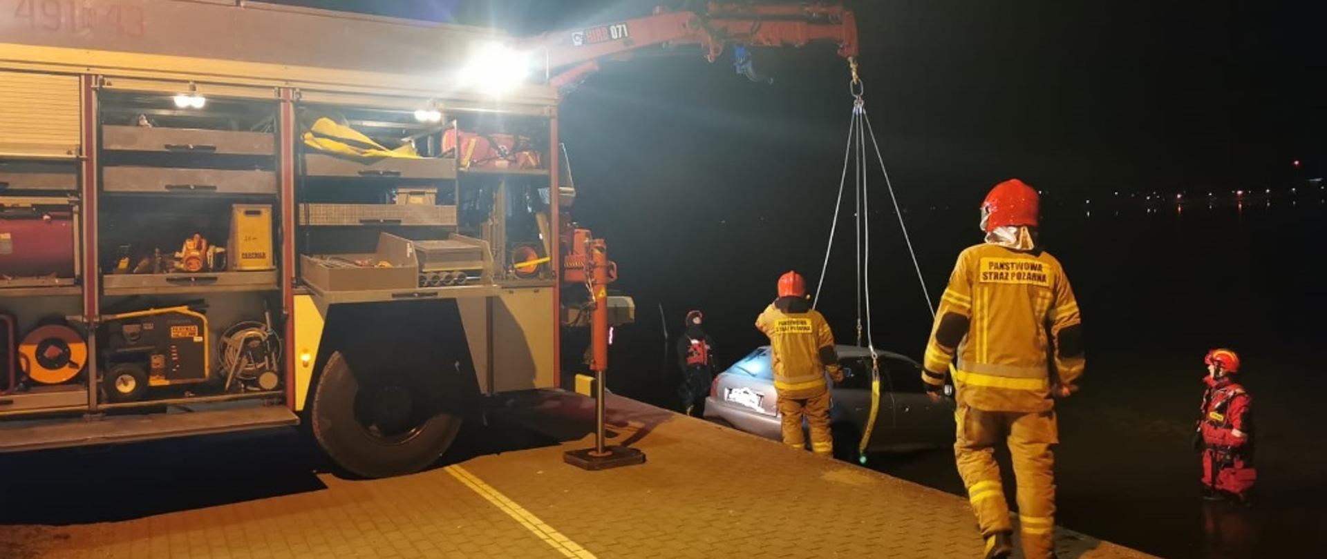 Straży podnoszą przy pomocy żurawia przewoźnego samochód na brzeg. 