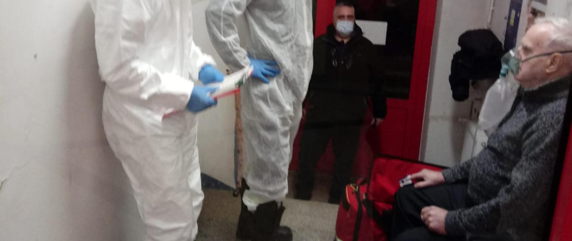 Dwóch strażaków w białych kombinezonach obok mężczyzna w masce tlenowej siedzący na krześle w tle mężczyzna ubrany na czarno w maseczce jednorazowej