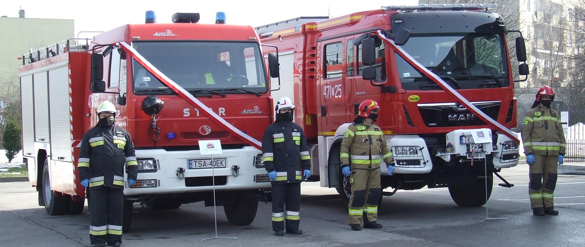 Zdjęcie zrobione na placu Komendy. Przedstawia dwa samochody strażackie. Na samochodach są zawieszone biało-czerwone szarfy. Przed każdym samochodem stoi dwóch strażaków, po prawej i po lewej stronie. Ubrani są w ubrania bojowe i hełmy a na twarzach mają ochronne maseczki.