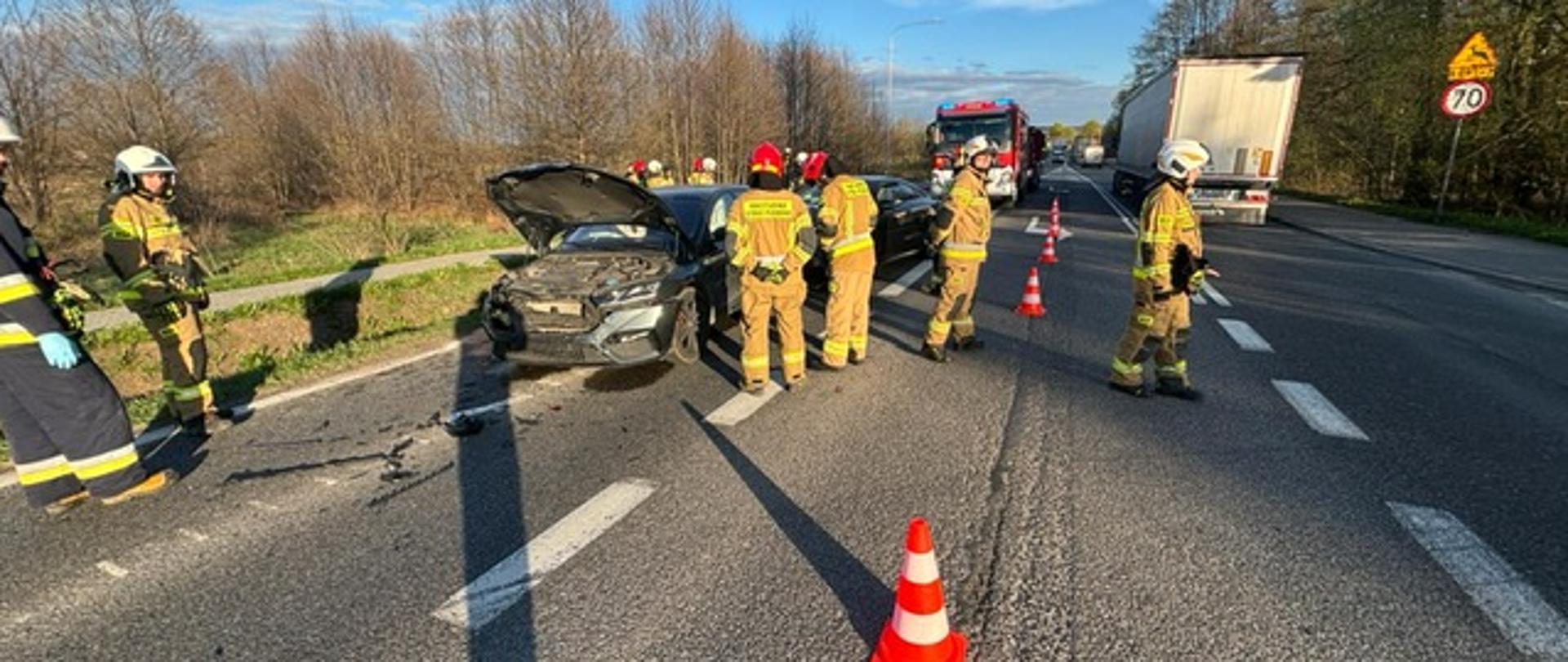 Zdjęcie przedstawia drogę krajową gdzie doszło do kolizji trzech aut. Po lewej stronie dwa z nich stoją na poboczu a przy nich ratownicy straży pożarnej. W głębi samochód pożarniczy, na jezdni ustawione stożki ostrzegawcze.