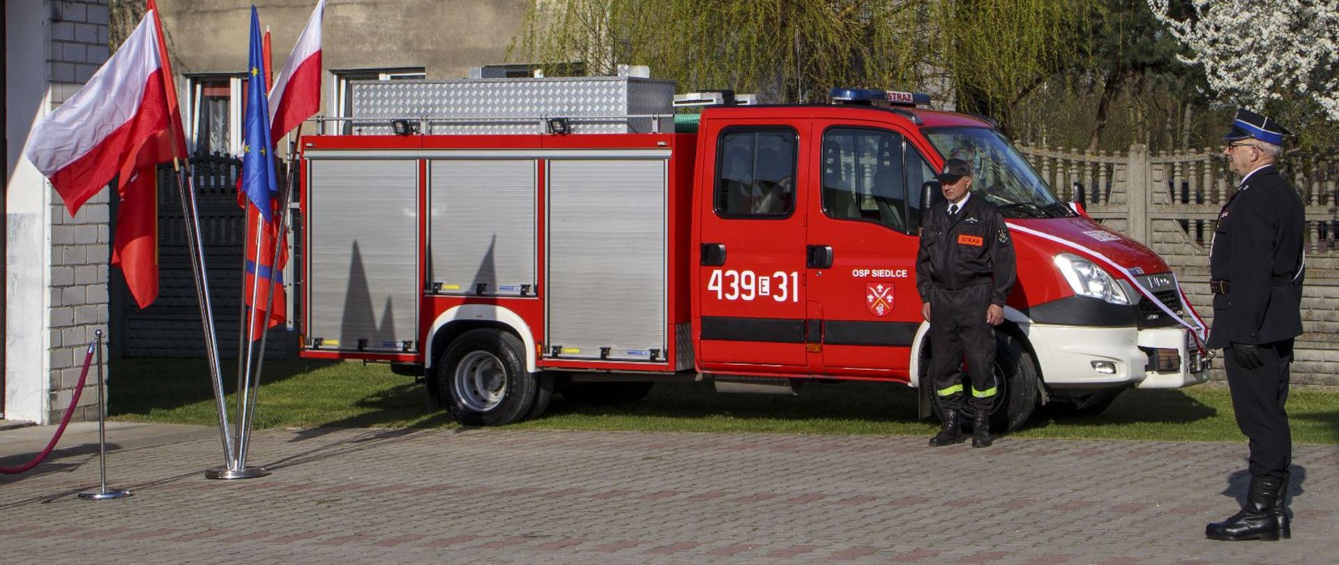 dwóch druhów osp stojących na placu, jeden w ubraniu koszarowych stoi przy samochodzie pożarniczym, drugi w umundurowaniu galowym stoi frontem bo budynku, w tle drzewa i budynek mieszkalny