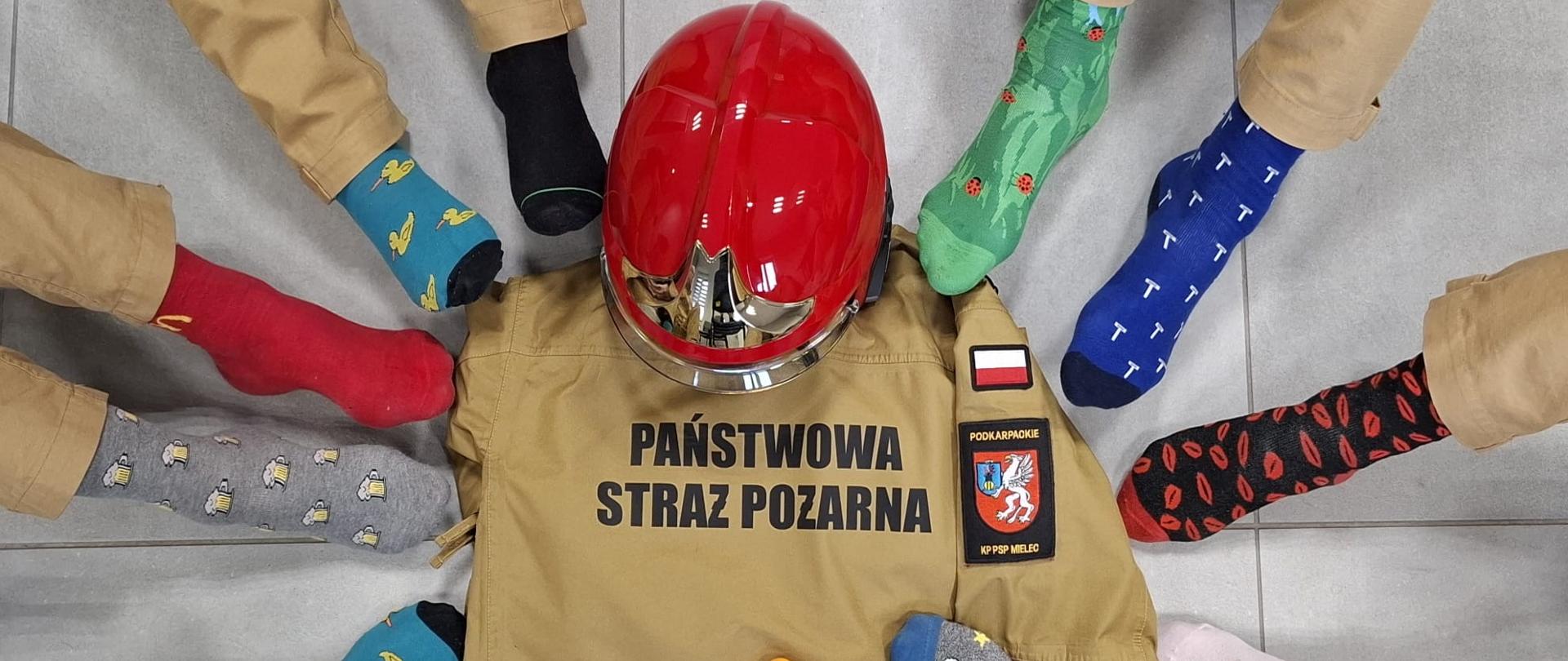 Widok z góry na okrąg utworzony przez stopy osób w nogawkach od piaskowych mundurów strażackich i kolorowych, niepasujących do siebie skarpetkach. W centrum okręgu leży kurtka mundurowa z napisem „PAŃSTWOWA STRAŻ POŻARNA” oraz czerwony hełm strażacki.