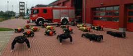 Zdjęcie strażaków wykonujących podpór przodem leżąc, na pierwszym planie czterech strażaków w umundurowaniu KDS, zaś na drugim planie 14 strażaków w umundurowaniu specjalnym. Wszyscy na planie budynku KP PSP w Brzezinach oraz samochodu bojowego z JRG Brzeziny 