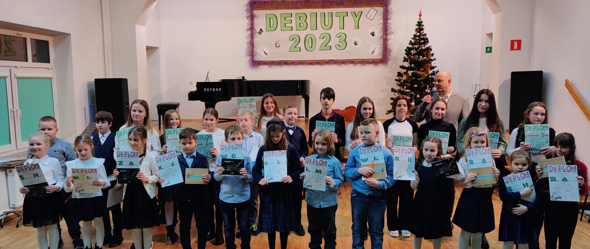 Zdjęcie pamiątkowe przedstawiające uczniów klas pierwszych po uroczystym pasowaniu w trakcie Koncertu Debiutów w auli szkoły.