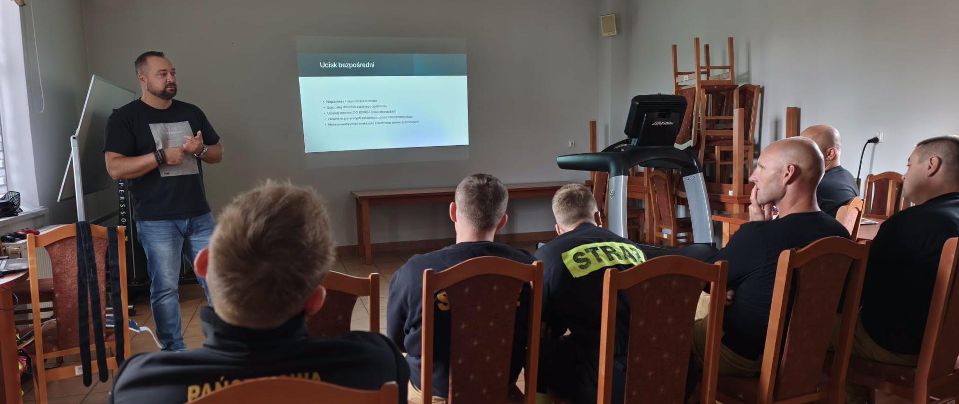 Sala wykładowa, z rzutnikiem i białymi ścianami. Instruktor w czarnym t-shircie i dżinsach stoi przed słuchaczami i prowadzi prezentację wyświetlaną z rzutnika na ścianie. Widok z tyłu na grupę strażaków siedzących w rzędach krzeseł i słuchających prelegenta.
