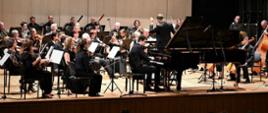 Zdjęcie przedstawiające pianistę - Ludwika Wiśniewskiego na scenie filharmonii. W tle orkiestra symfoniczna. 