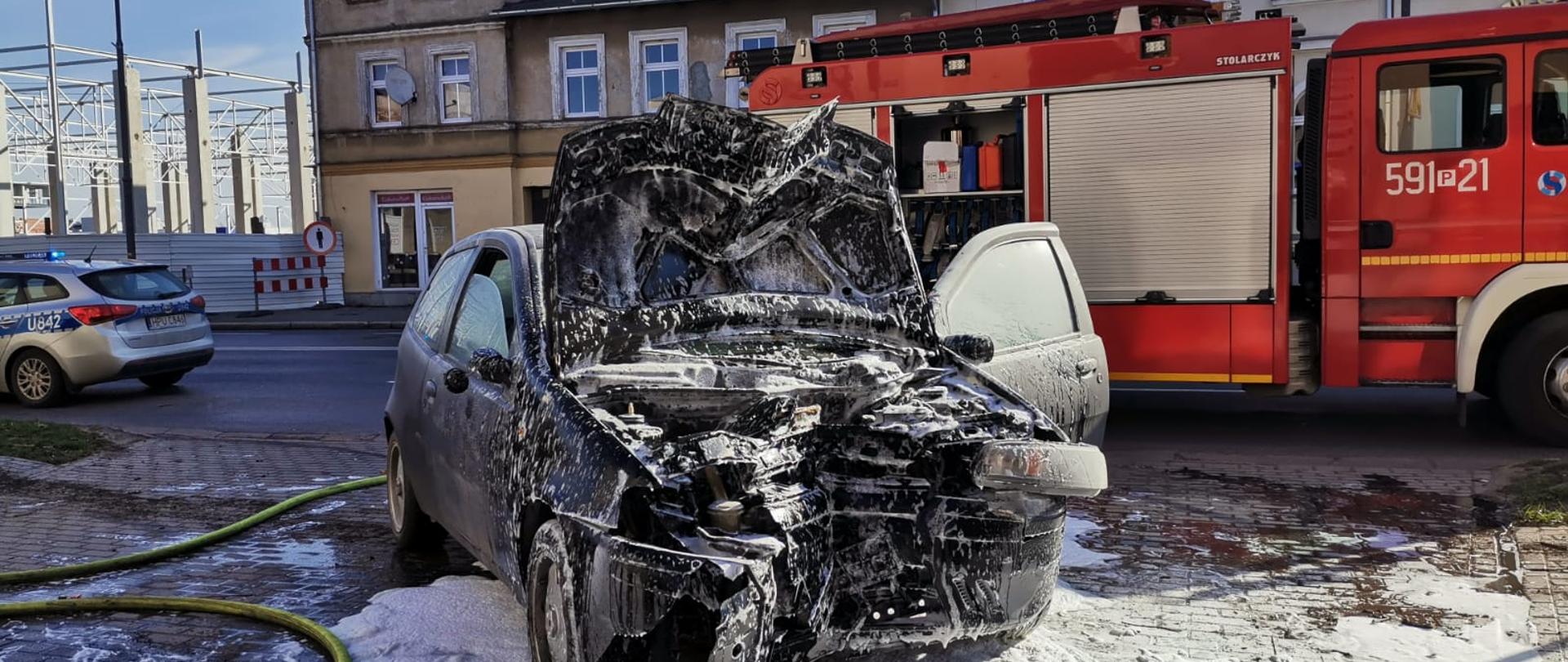 Widać uszkodzony przód samochodu, w koło znajduje się piana gaśnicza, po lewej samochód Policji, z tyłu samochód Straży Pożarnej