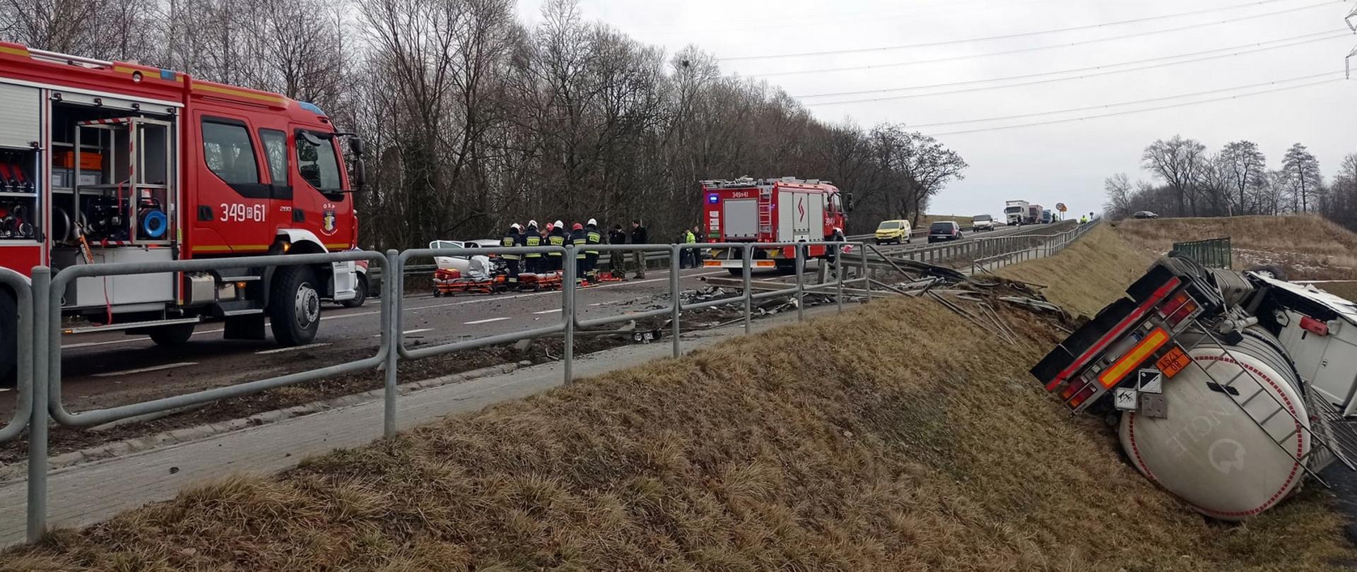 Zdjęcie zrobione na zewnątrz. Po prawej stronie zdjęcia na skarpie drogowej leży wywrócony samochód cysterna. Na jezdni stoją dwa samochody pożarnicze. W tle, na jezdni stoi rozbity samochód osobowy, a przy nim strażacy prowadzący działania ratownicze.
