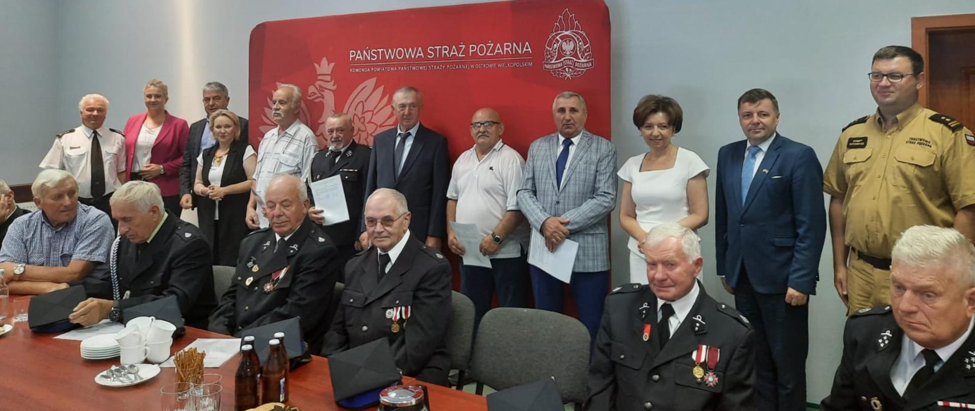 zdjęcie grupowe, wśród osób stojących i siedzących sa strażacy , w tle czerwona ścianka z orłem