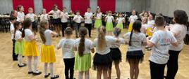 Zdjęcie przedstawia grupę uczniów tańczących w okręgu na sali koncertowej. Uczniowie są ubrani w czarne spodnie i białe koszulki, uczennice mają zielone i źółte spódniczki.