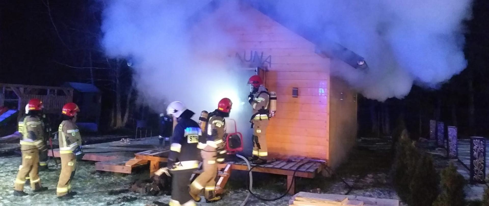 Zdjęcie przedstawia drewniany budynek sauny, z którego wydobywa się biały dym. Koło budynku widać kilku strażaków w umundurowaniu specjalnym, którzy prowadza działania gaśnicze. Zdjęcie zrobione w porze nocnej, na ziemi zalega warstwa białego śniegu.