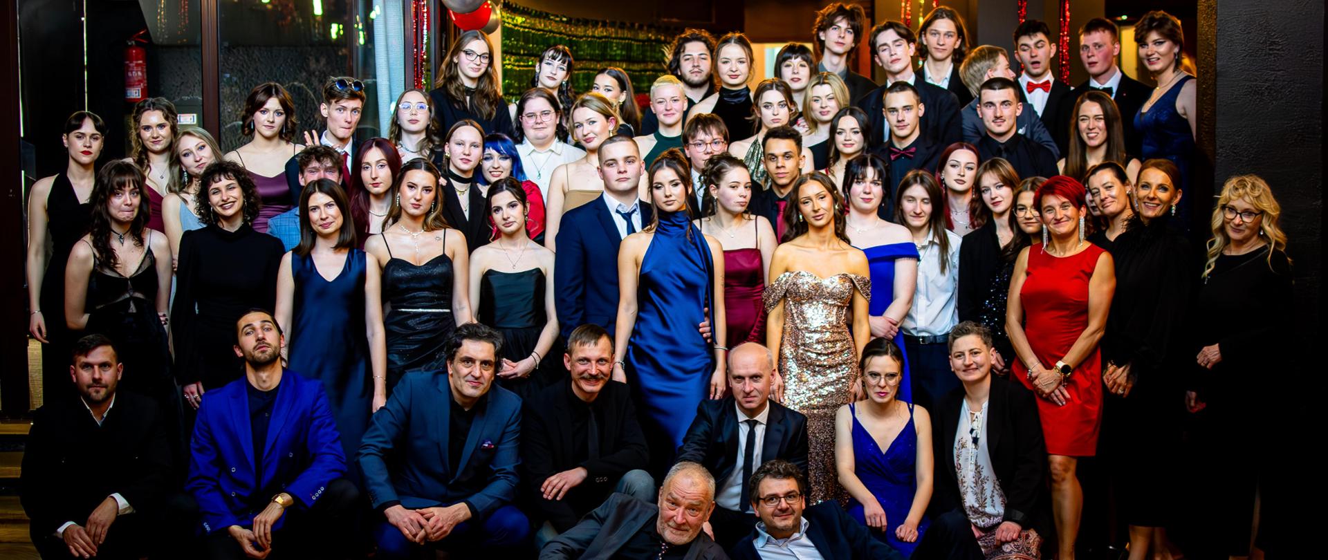 Na zdjęciu widzimy uczniów klas maturalnych, nauczycieli, wychowawców i dyrekcję szkoły. Panie i panowie ubrani są odświętnie w stroje wieczorowe. Na pierwszym planie widzimy dwóch wychowawców leżących na parkietowej podłodze klas: 5A i 5B, jest to pan Piotr Bies i pan Bartłomiej Marduła. W drugim rzędzie nauczycieli kucają do zdjęcia. Od lewej strony: Wojciech Bachleda-Dorcarz, Adam Soroczyński, Artur Kapturski, Tomasz Szybka, Marek Król-Józaga dyrektor szkoły, Paulina Siedlarz. Kolejny plan to uczennice i uczniowie klas piątych, a wśród nich: Anna Gąsienica-Roj, Agnieszka Czajkowska wicedyrektor szkoły, Izabela Gąsior, Aleksandra Karpińska i Magdalena Bilczewska-Wiśniewska. Na trzecim planie widzimy ciemne ściany i okna udekorowane w srebrne i czerwone baloniki.