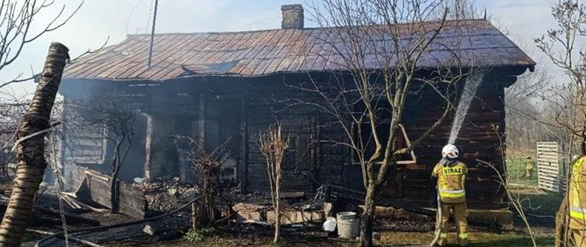 Na zdjęciu widoczny jest dom drewniany pokryty starą zardzewiałą i częściowo przegrzaną blachą. Na środku dachu znajduje się komin natomiast z lewej strony z dachu wystaje metalowa rura od przyłącza elektrycznego. Front budynku częściowo spalony. Spalone drzwi i okno. Przed budynkiem leżą kawałki spalonych desek oraz nadpalone meble z wnętrza budynku. Przed budynkiem cztery małe drzewa bez liści. Podłoże przed budynkiem nieutwardzone pokryte w całości trawa na której leży linia gaśnicza. Z prawej strony widocznych dwóch strażaków odwróconych plecami. Ubrani są w ubrania specjalne w kolorze piaskowym i białe hełmy. Jeden z nich, stojący przy budynku trzyma w rękach prądownice wodną i podaje rozproszony prąd wody na budynek. Z prawej strony za budynkiem widoczna biała brama wjazdowa na posesje oraz jeden strażak.