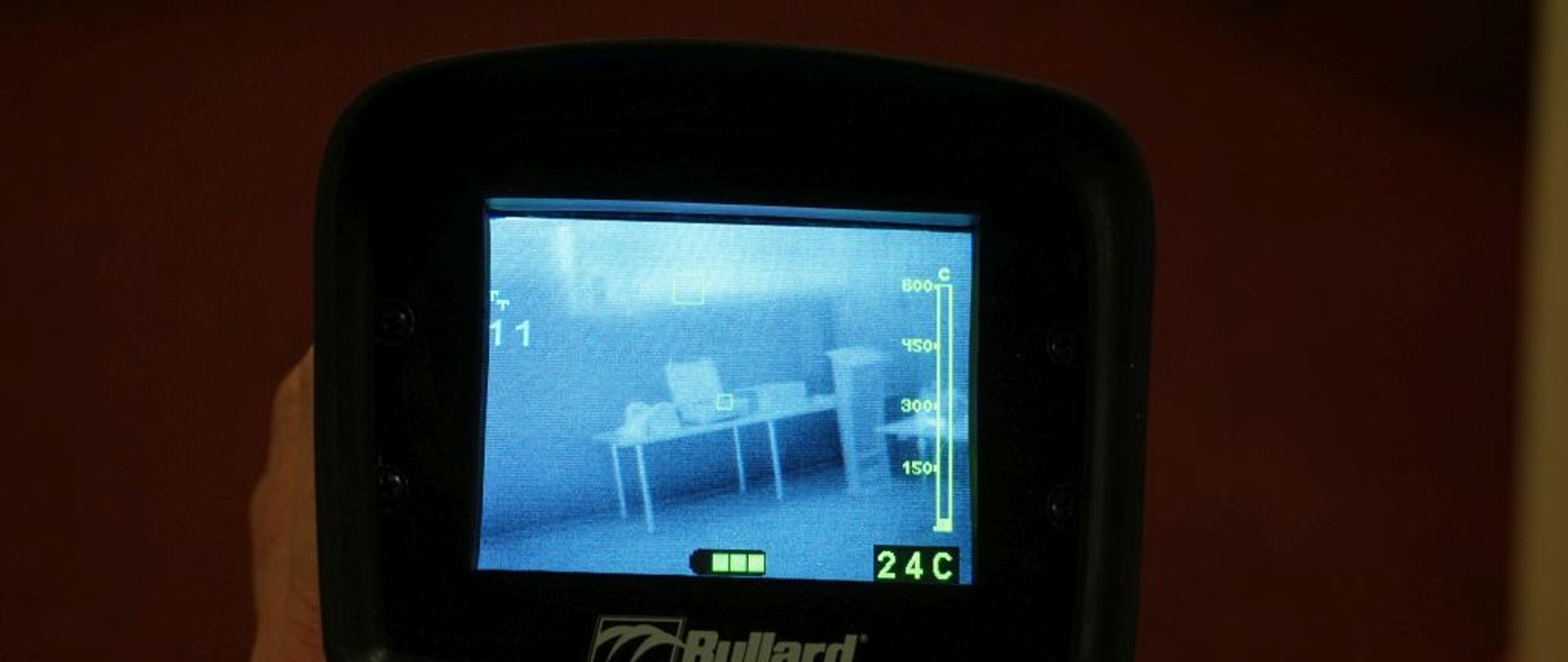 Na wyświetlaczu kamery termowizyjnej widać biurko oraz okno. Obok skala z temperaturą.