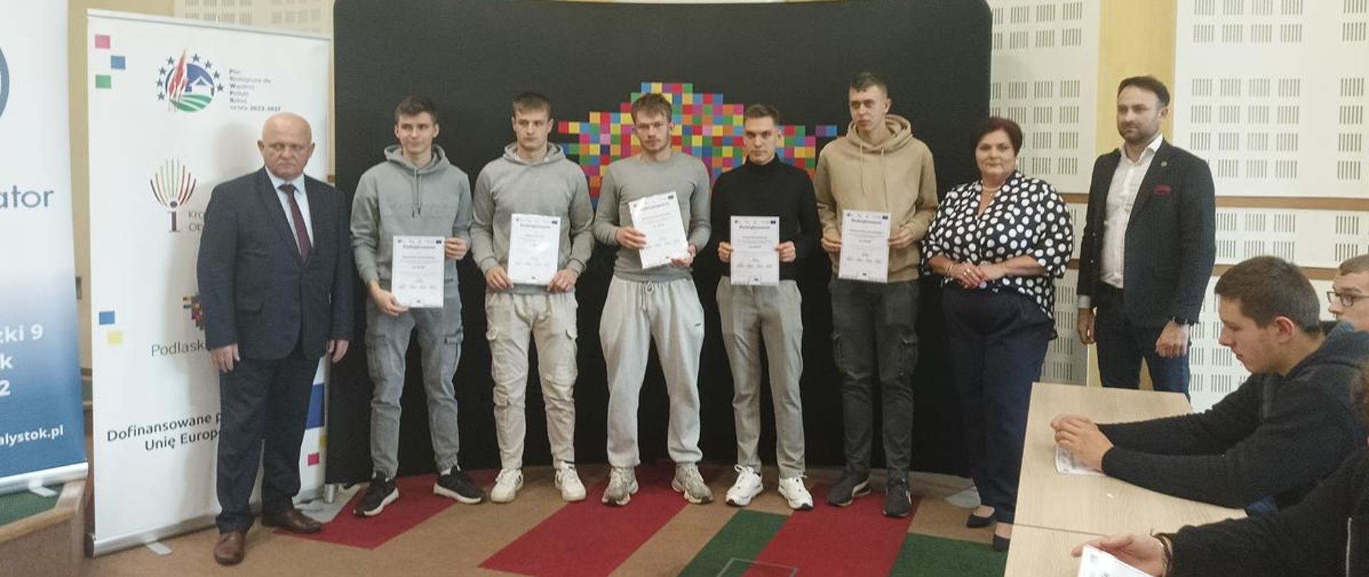 Grupa pięciu uczestników konkursu prezentuje otrzymane dyplomy, stojąc pomiędzy dwiema osobami w strojach biznesowych oraz kobietą w czarno-białej bluzce. Uroczystość odbywa się w jasnej sali konferencyjnej.