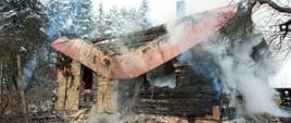 Pożar drewnianego budynku mieszkalnego. Budynek po przejściu pożaru z otworów okiennych drzwiowych i spod dachu wydobywa się dym.