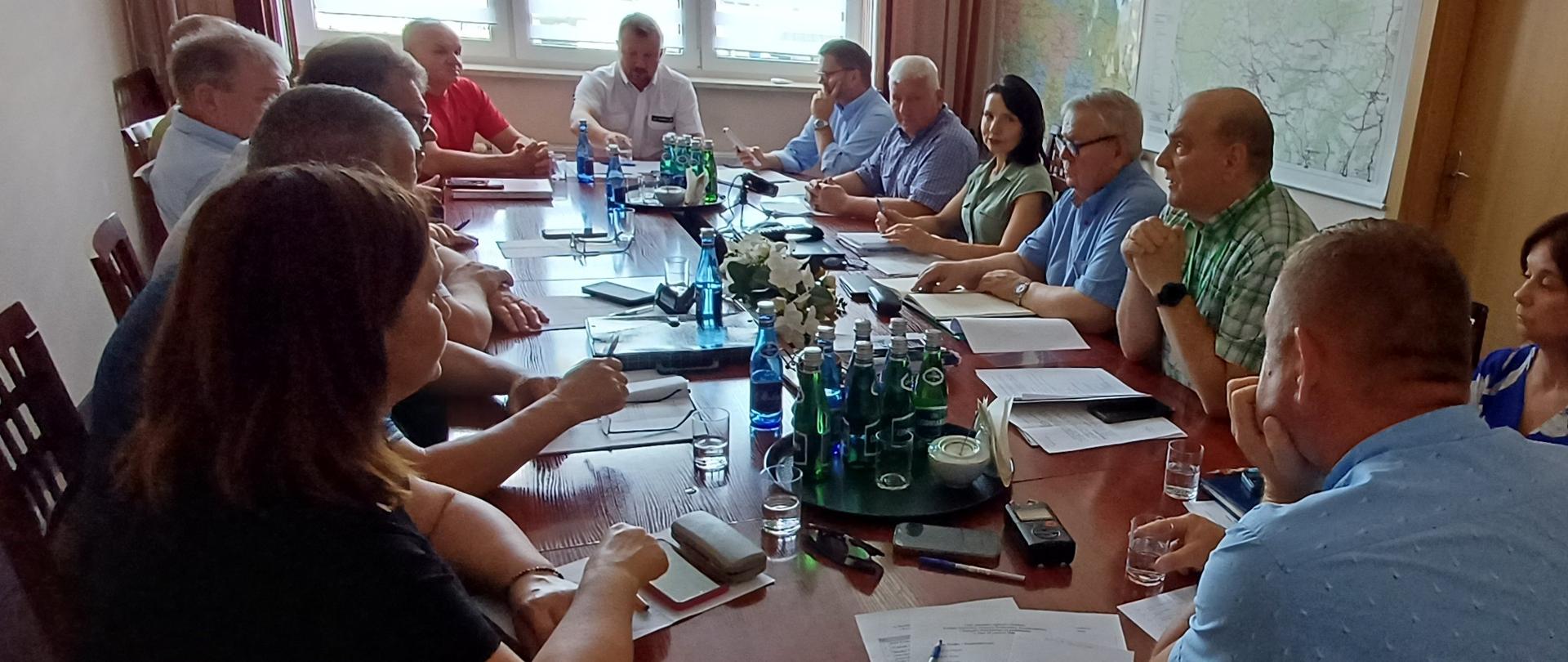 W sali konferencyjnej odbywa się spotkanie kilkunastu osób siedzących wokół długiego, drewnianego stołu. Uczestnicy dyskutują nad dokumentami rozłożonymi przed nimi. Na stole znajdują się kartki, notatniki, długopisy, telefony komórkowe, szklanki z wodą oraz kilka butelek wody mineralnej ustawionych na okrągłych tacach. W centralnej części stołu ustawiono niewielką kompozycję z białych kwiatów.
Na końcu stołu siedzi mężczyzna w białej koszuli, który sprawia wrażenie prowadzącego spotkanie. Pozostali uczestnicy zwracają uwagę w jego stronę lub prowadzą rozmowę z osobami siedzącymi naprzeciwko. Po prawej stronie stołu siedzą między innymi kobieta w zielonej bluzce oraz mężczyzna w zielonej koszuli w kratę, który wydaje się zabierać głos. W pomieszczeniu znajduje się około piętnastu osób – kobiet i mężczyzn.
W tle widoczne są duże okna z roletami przepuszczającymi światło dzienne, zasłony oraz duża mapa zawieszona na ścianie. Zdjęcie przedstawia formalne posiedzenie lub naradę w urzędzie albo instytucji, podczas której omawiane są dokumenty i prowadzone są dyskusje.